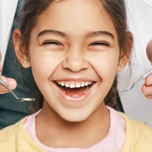 A smiling child with open mouth, surrounded by dental tools held by a dentist, wearing a yellow and pink outfit.