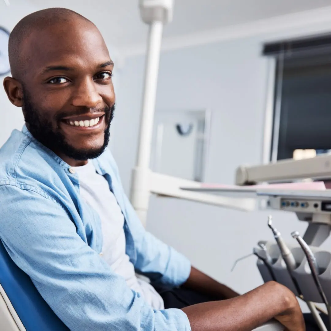 A person is sitting in a dental chair, smiling at the camera. The background shows dental equipment and a brightly lit room.