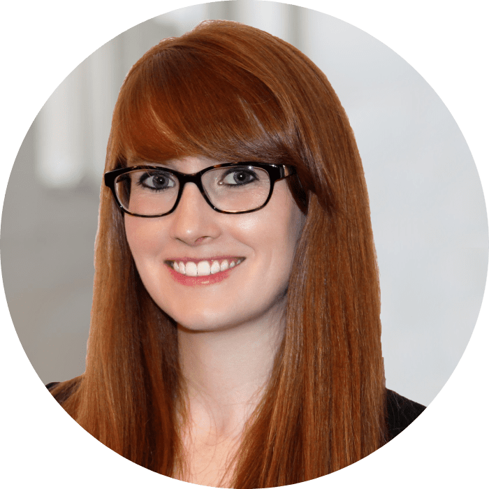 A person with long red hair and glasses smiles at the camera against a neutral background.